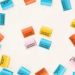 Bunte Weblabels mit Botschaften wie „I AM KIND" und „I AM LOVED" verstreut auf hellem Untergrund.