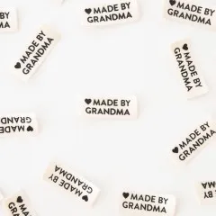 Nahaufnahme mehrerer „Made by Grandma"-Weblabels in Creme und Schwarz, verstreut auf weißem Untergrund.