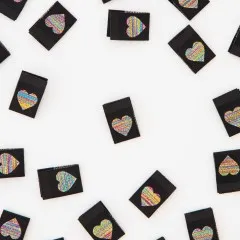 Nahaufnahme mehrerer schwarzer Weblabels mit glänzend besticktem Regenbogen-Herz-Motiv, verstreut angeordnet.