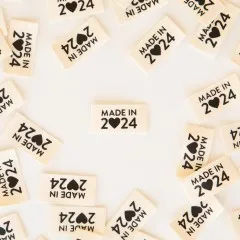 Naturfarbene Weblabels mit Schriftzug „Made in 2024" und Herz-Symbol verstreut auf hellem Untergrund.