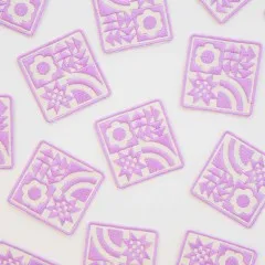 Mehrere Lavender-Quilt-Block-Aufnäher locker verstreut auf weißem Untergrund.