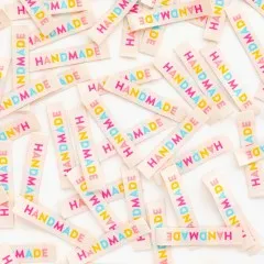 Dichte Nahaufnahme vieler verstreuter „HANDMADE"-Weblabels in Regenbogenfarben auf Creme.