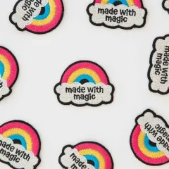 Mehrere Regenbogen-Patches „Made with Magic" verstreut, teils auf dem Kopf liegend.