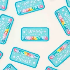Nahaufnahme mehrerer türkisfarbener „Sewing is my Superpower"-Patches, dicht verstreut.