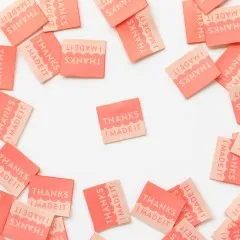 Mehrere korallenfarbene Sarah-Hearts-Weblabels „Thanks I Made It" in Koralle und Creme, ein Label mittig freigestellt.