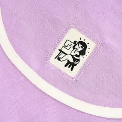 Ein Label mit einer an der Nähmaschine sitzenden Figur ist diagonal am Saum eines lilafarbenen Kleidungsstücks mit cremefarbener Paspel angenäht.