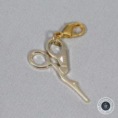 Nahaufnahme des goldenen Storchenscheren-Anhängers mit Karabiner auf grauem Stoffuntergrund.