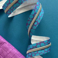 Nähere Ansicht des Blue-Pebble-Reißverschlussbands, das sich über die Kante eines pink-violett gemusterten Stoffs schlängelt.