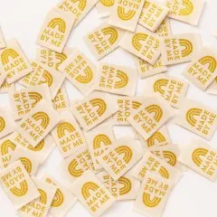 Dicht verstreute cremefarbene Weblabels mit goldenem Regenbogen-Motiv "Made by Me" in Nahaufnahme.