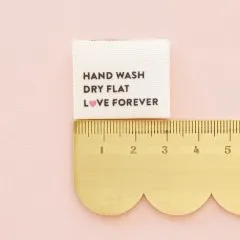 Einzelnes „Hand Wash, Dry Flat, Love Forever“-Label neben einem goldenen Lineal zum Größenvergleich.