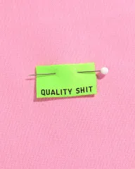 Neongrünes 'Quality Shit'-Label mit Stecknadel auf rosa Stoff