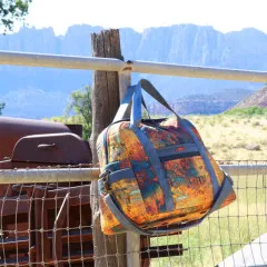 Vintage-Print-Reisetasche hängt an einem Weidezaun vor Felsenkulisse im Freien.
