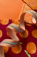 Orange-weiß gestreiftes Reißverschlussband drapiert über Stoffen in Orange-Lederoptik, grafischem Rautenmuster und dunkelrotem Stoff mit Fruchtmotiv.