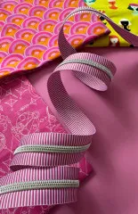 Pink-lila gestreiftes Reißverschlussband drapiert über bunten Stoffen mit Regenbogen-Fächermuster, gepunktetem Muster und einem gelben Stoff mit Figurenmotiv.