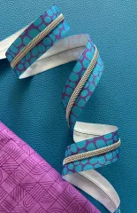 Blau-violett gemustertes Pebble-Reißverschlussband geschwungen auf türkisfarbenem Lederimitat-Untergrund neben pink-violett gemustertem Patchworkstoff.