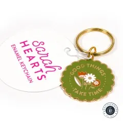 Sarah Hearts Emaille-Schlüsselanhänger „Good Things Take Time“ mit Blumenmotiv, neben der Anhängekarte.