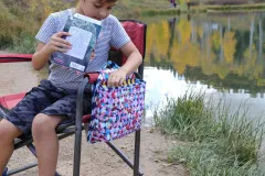 Kind greift in die Sidekick-Satteltasche in Konfetti-Print, befestigt an einem Campingstuhl am See.