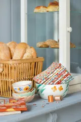 Gefaltete Tilda-Blenderstoffe der Kollektion Village Life in einer Keramikschale, dekoriert neben einem Brotkorb und Garnrollen.