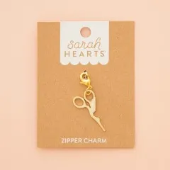 Zipper Charm in Form einer Storchenschere auf Verpackungskarte, fotografiert vor zartrosa Hintergrund.