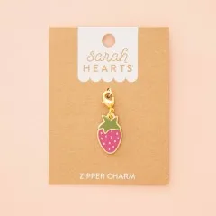 Erdbeer-Zipper-Charm auf Verpackungskarte, fotografiert vor zartrosa Hintergrund.