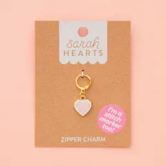 Zipper Charm auf Verpackungskarte, fotografiert vor zartrosa Hintergrund.