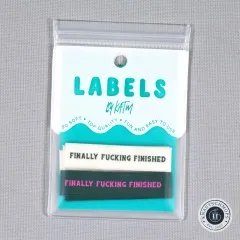 Verpackungskarte „Labels by KATM" mit den „Finally Fucking Finished"-Labels in Creme und Waldgrün, gerade von vorne fotografiert.