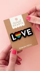 Verpackungskarte des Love-Pride-Patches in der Hand gehalten, frontal fotografiert vor rosa Hintergrund.