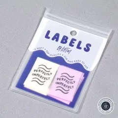 „Perfectly Imperfect"-Labels in Elfenbein und Flieder, sichtbar in der Original-Verpackungskarte „Labels by KATM", schräg fotografiert.