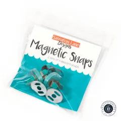 Originalverpackung der Sassafras Lane Magnetic Snaps mit dem Hinweis „2 sets of iridescent snaps", schräg fotografiert mit Blick auf die türkisfarbenen Druckknopf-Teile.