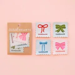Verpackung neben vier Handmade-Weblabels mit Schleifenmotiv in Rot, Grün, Blau und Pink.