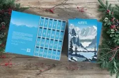 Geöffneter Adventskalender neben zwei geschlossenen Verpackungen, dekoriert mit Tannenzweigen und roten Beeren auf Holzuntergrund.