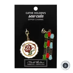 Zwei goldene Emaille-Anhänger von Cathe Holden: ein Stickrahmen mit Rosen-Stickmotiv und drei Stickgarn-Anhänger mit Aufschrift 'Stitch', auf Verpackungskarte.