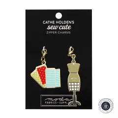 Zwei goldene Emaille-Anhänger von Cathe Holden: ein Stapel gemusterter Stoffballen und eine Schneiderpuppe, auf Verpackungskarte.