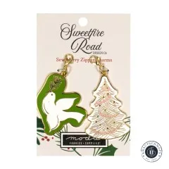 Zwei goldene Emaille-Anhänger von Sweetfire Road: ein bestickter weißer Weihnachtsbaum und eine grün-weiße Friedenstaube mit Tannenzweig, auf Verpackungskarte 'Sew Merry Zipper Charms'.